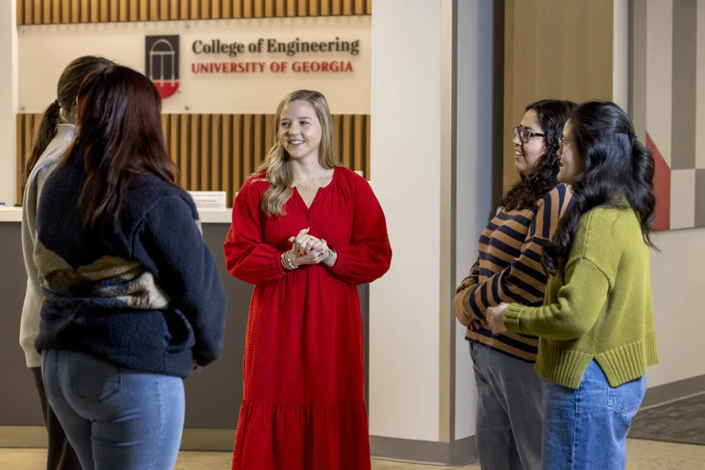 A woman stands in the hallway of an academic building, having a conversation with the students around her.