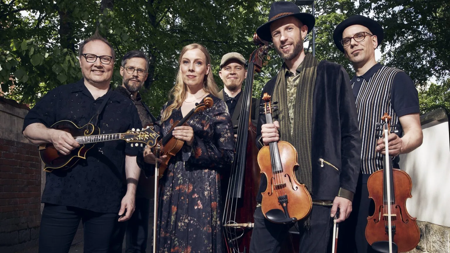A band consisting of five men and one woman stands outside, holding their instruments and gazing at the camera.