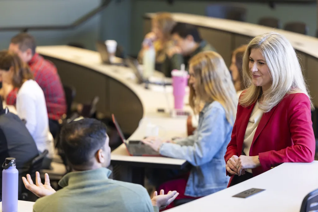 A woman on the right in the foreground talks with a man sitting near her in large classroom with others silhouetted behind them.