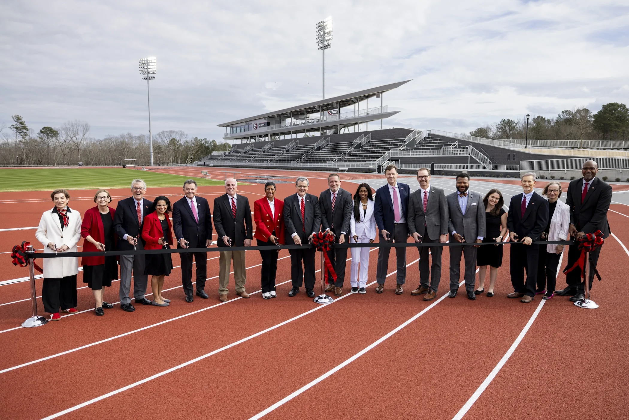 A group of university officials and athletic leaders hold ceremonial scissors along a black ribbon stretched across a red running track during a facility dedication, with a modern grandstand and track lanes extending into the background.