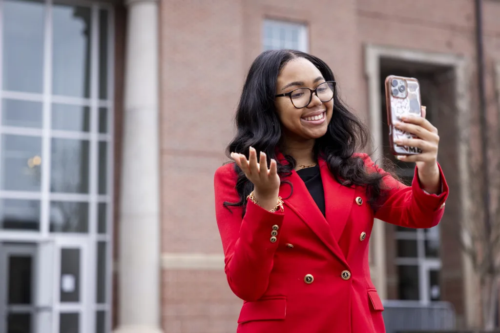 A woman looks into the camera of her phone, filming a video for social media, outside an academic building.