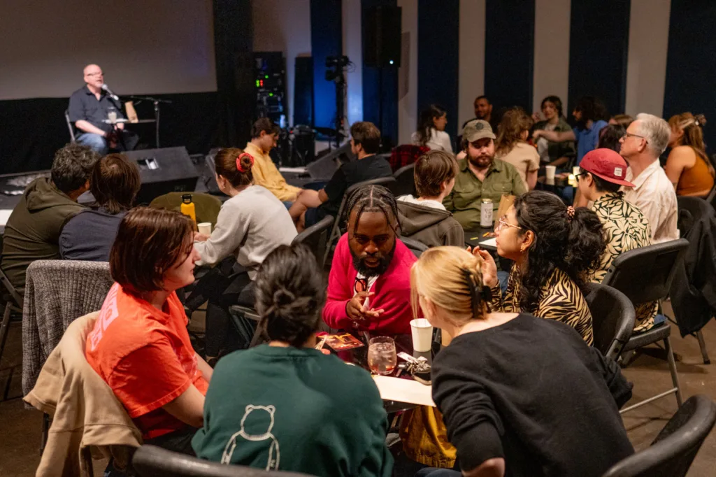 Audience members sit around small tables in a dimly lit venue during a trivia-style event while a speaker sits at a microphone onstage in the background.