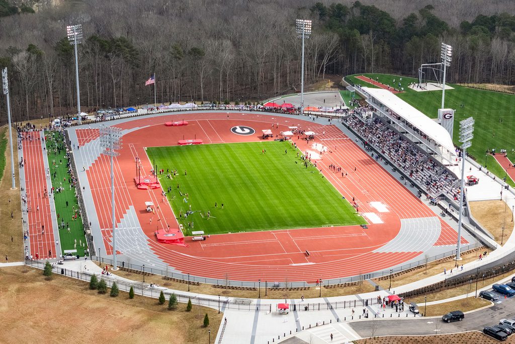 A wide aerial photo shows a red oval track encircling a green field, with athletes, spectators and stands spread across the complex and trees lining the perimeter.