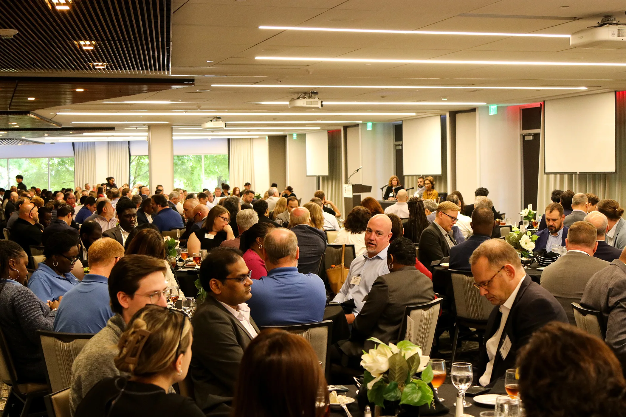 A large audience of attendees sits at round banquet tables in a conference room, listening to speakers at the front of the room.
