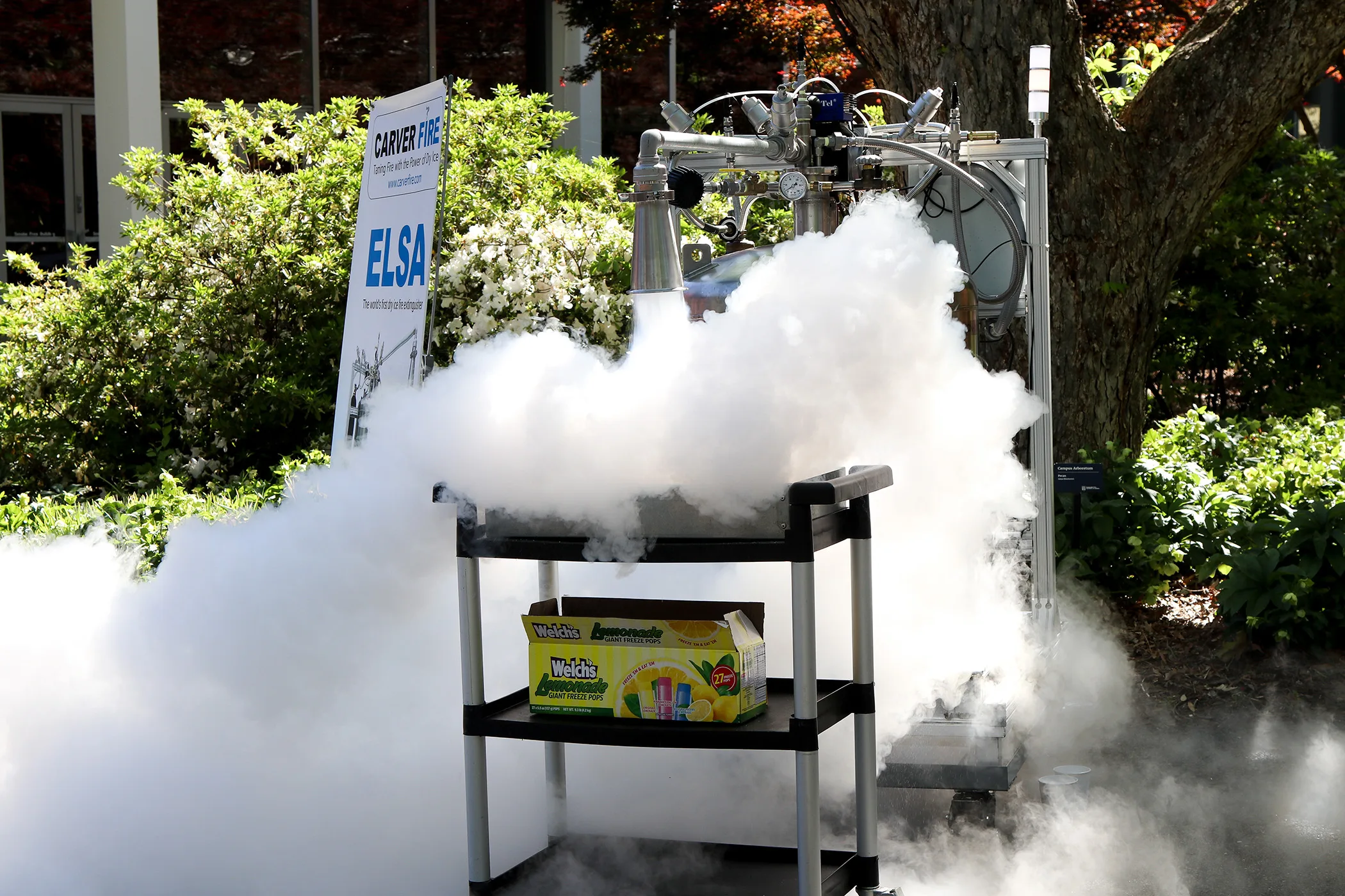 A liquid nitrogen fire suppression system releases thick white vapor beside a sign labeled “ELSA,” during a live outdoor demonstration.