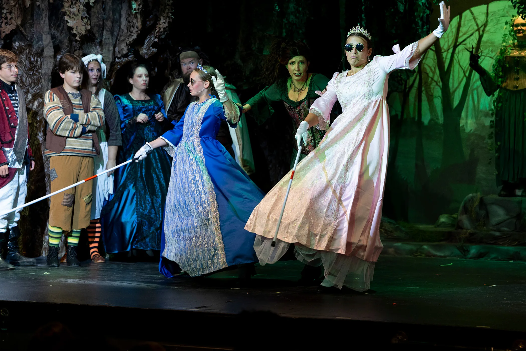 Actors in colorful costumes perform a scene onstage during a theatrical production, with a forest-themed set and dramatic lighting.