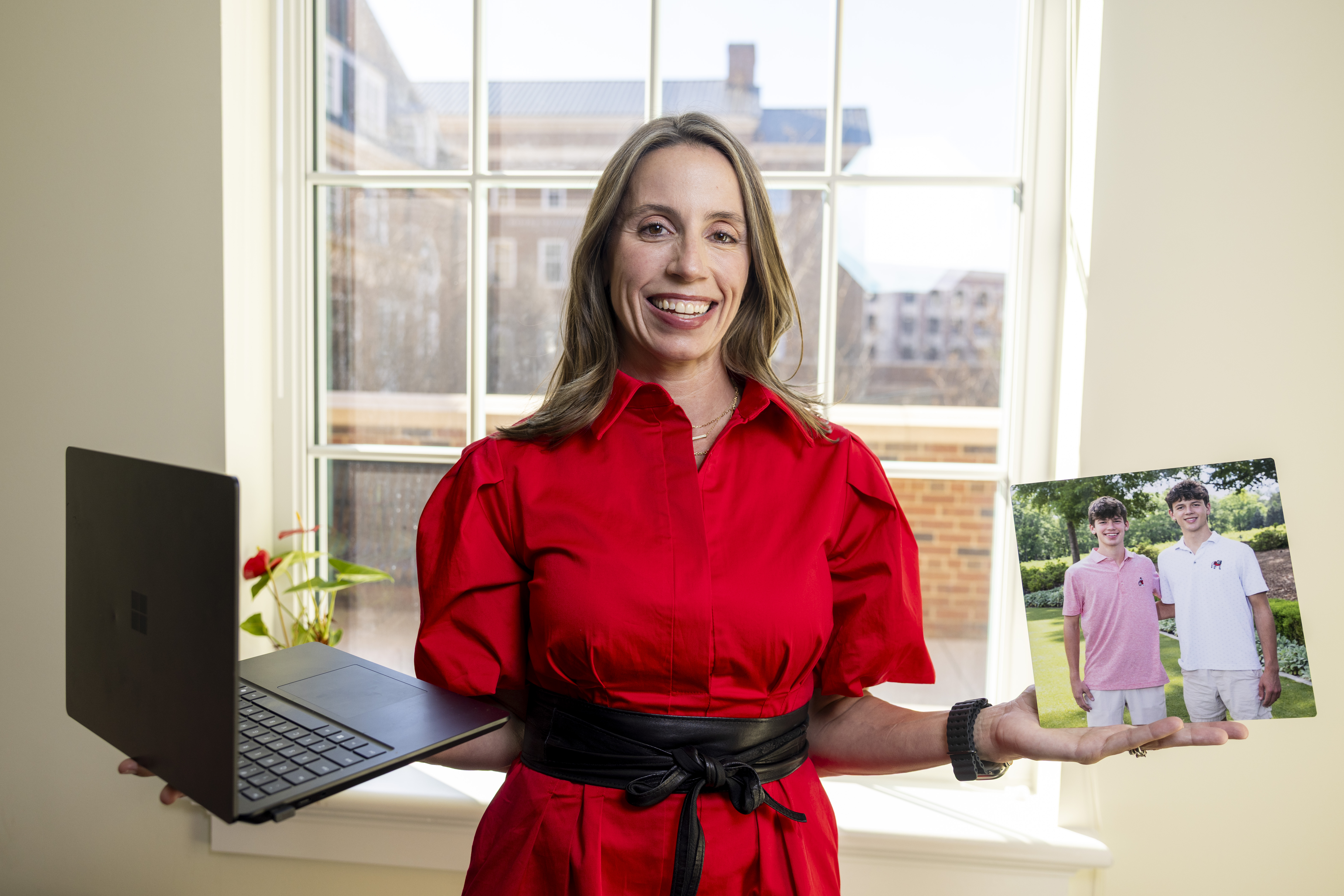 A woman in a red top holds a laptop in one hand and a photo of two boys in the other hand.