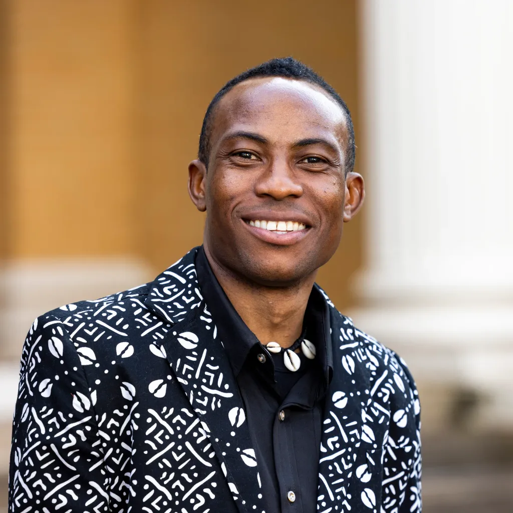 A closeup of a man in a black and white printed jacket smiling outside.