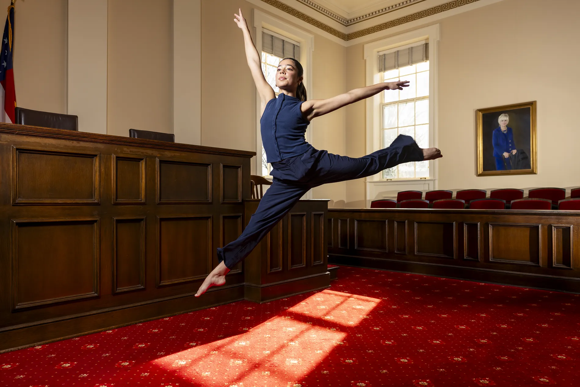 A female dancer in a dark blue top and dark blue pants leaps into the air.