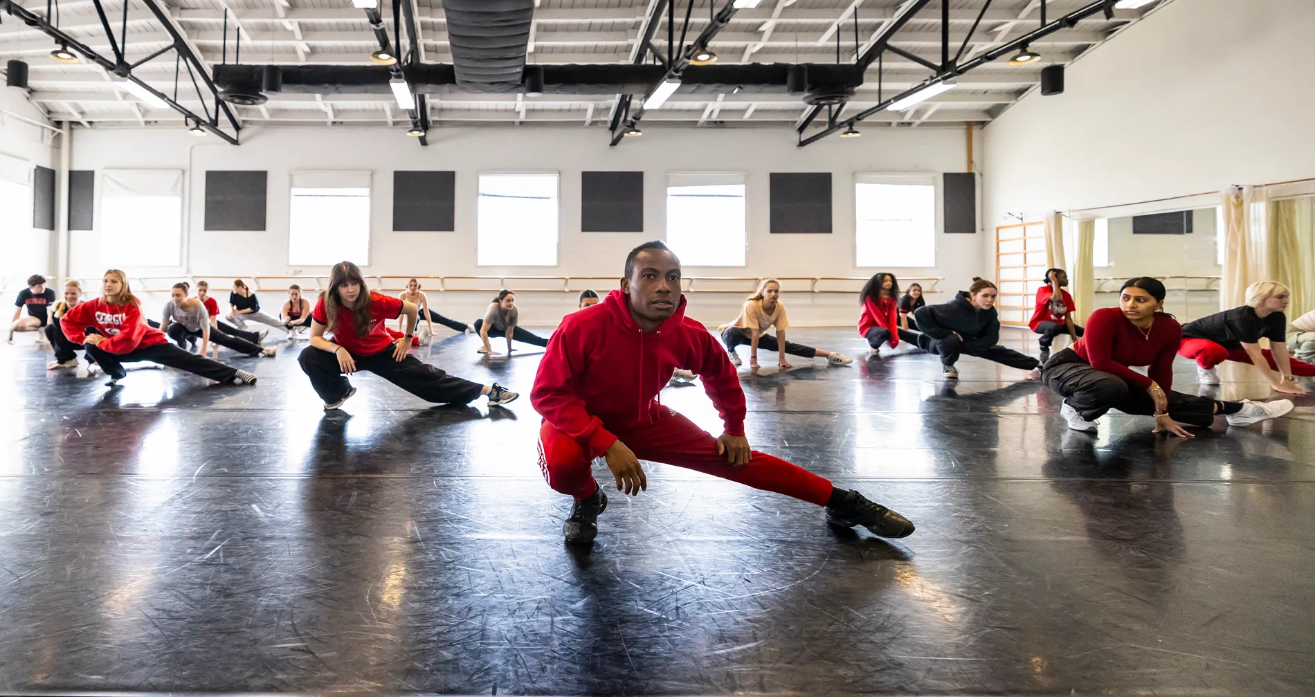 Several dancers in a studio crouch into a position with one leg bent at the knee and the other leg straight out to the side.