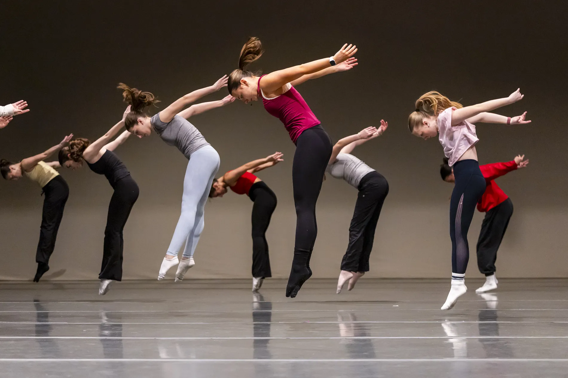 A group of dancers in a studio jump into the air, looking down with their arms stretched behind them.