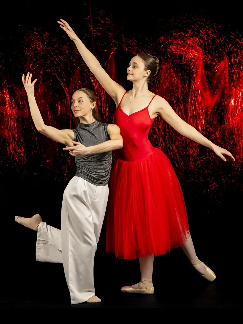 Two female dancers pose together in front of a red and black background.