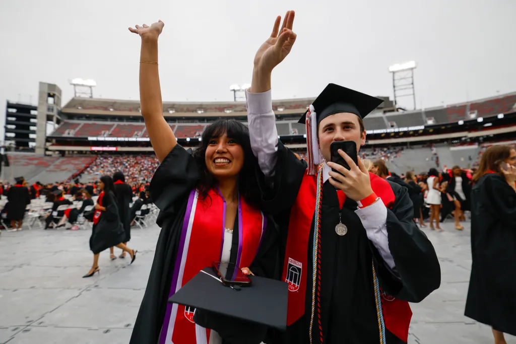 Two graduates in caps and gowns raise their hands to wave, one holding a phone as they look toward the stands of the stadium.