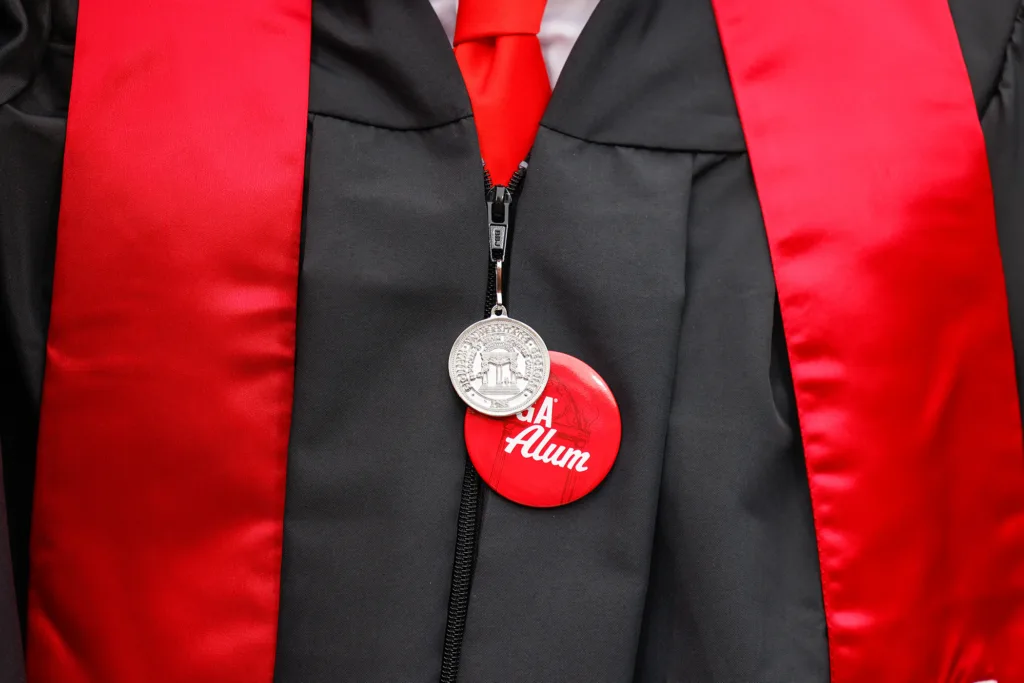 A close-up of a graduation gown shows a red button that reads “UGA Alum” pinned to the front.