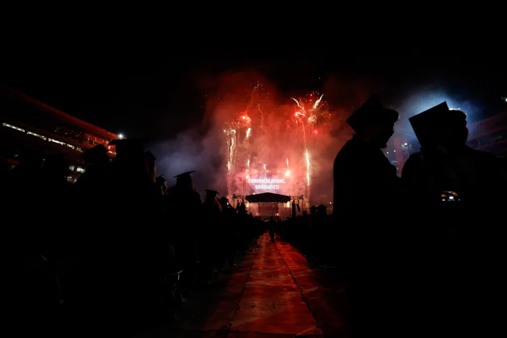 Silhouetted graduates stand facing the stage as fireworks explode overhead, with a screen in the distance reading “Congratulations, Graduates!”