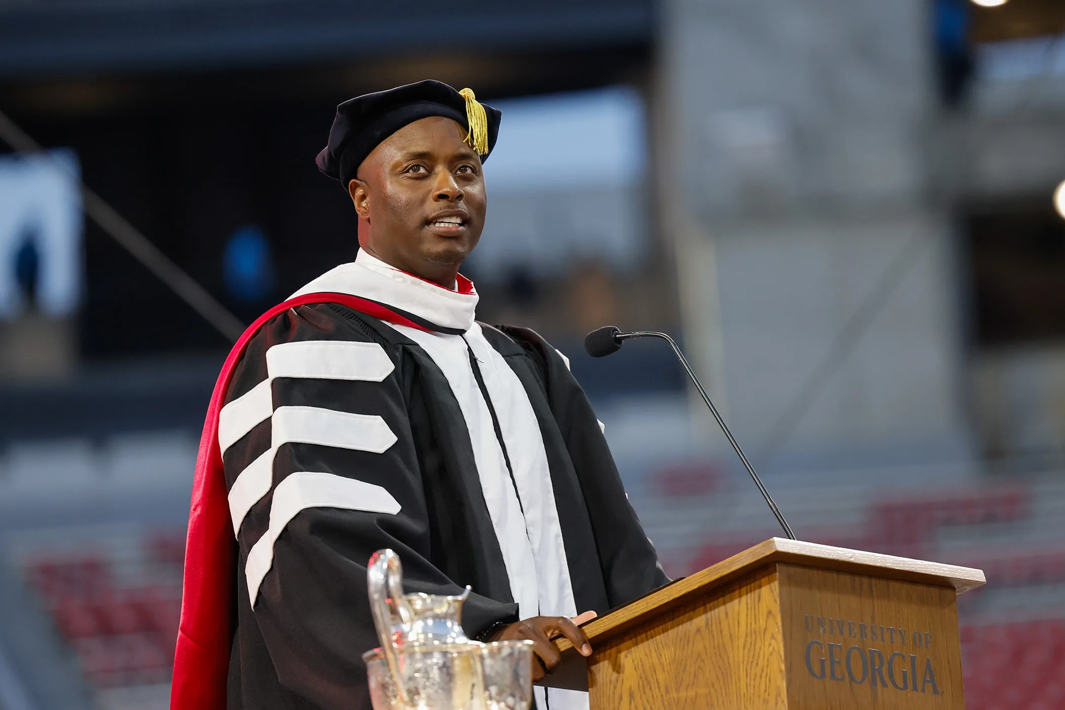 DJ Shockley in regalia stands at a podium, addressing the Commencement crowd inside the stadium.