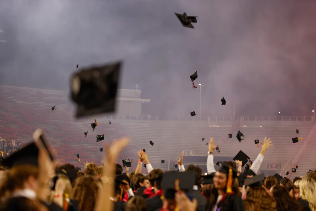 Graduates toss their caps into the air as hands reach up in celebration across a crowded stadium floor.