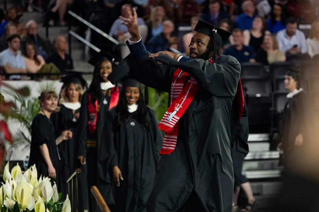 A graduate in a black gown and red stole gestures playfully while walking across the stage, with other graduates lined up behind him.