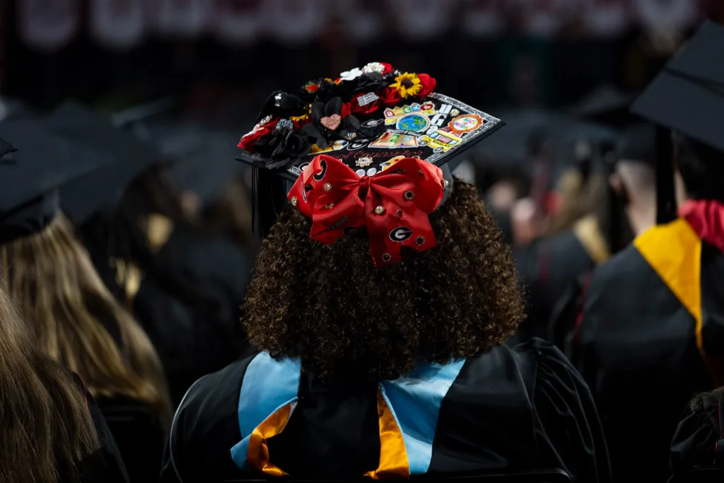 The back of a graduate’s decorated cap features flowers, stickers and a large red bow, standing out among a crowd of seated graduates.