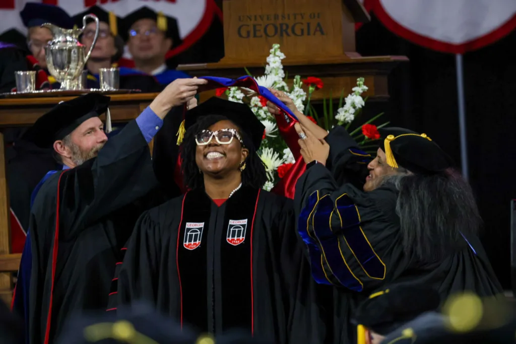 A smiling graduate in cap and gown is hooded on stage by faculty members during the doctoral Commencement ceremony.