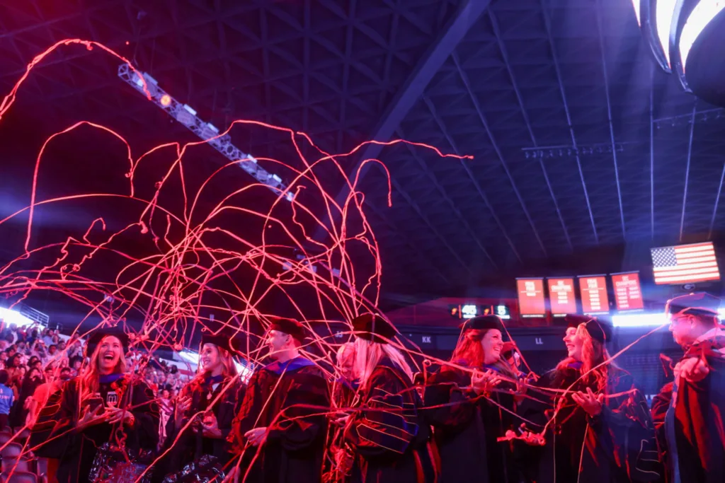 Red streamers arc through the air over graduates celebrating under the bright lights.