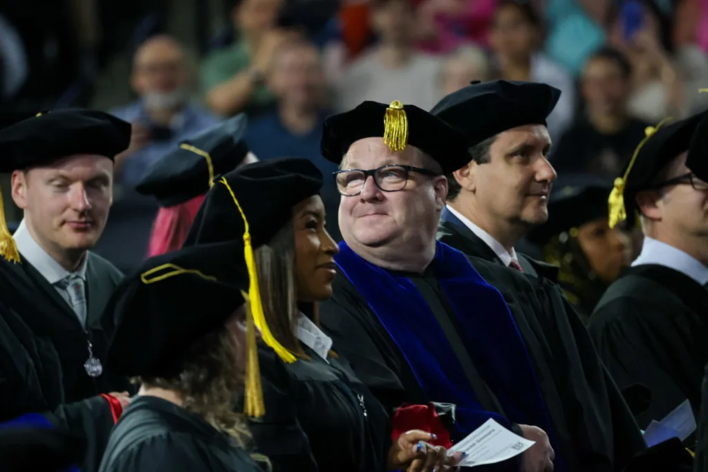 People in academic regalia sit in a row, watching the doctoral Commencement ceremony with attentive expressions.