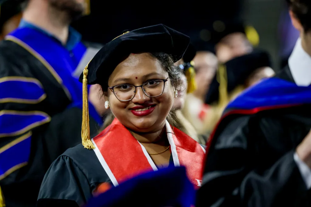 A doctoral candidate wearing glasses and a red stole smiles toward the camera while standing among fellow graduates.