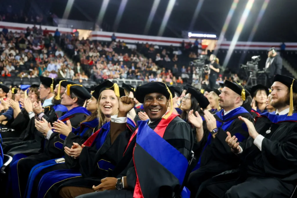Seated graduates in academic regalia clap and cheer, with one raising a fist in celebration as the crowd looks on.