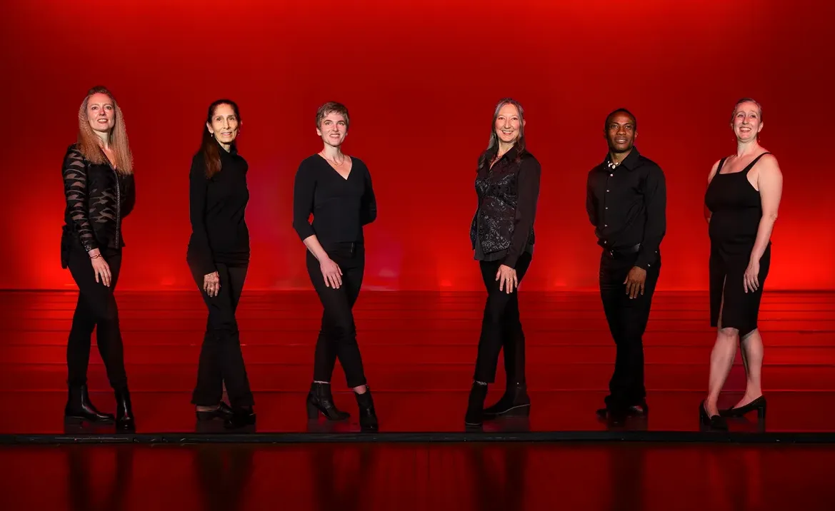 A group of women in black clothing pose on a stage in front of a red background.
