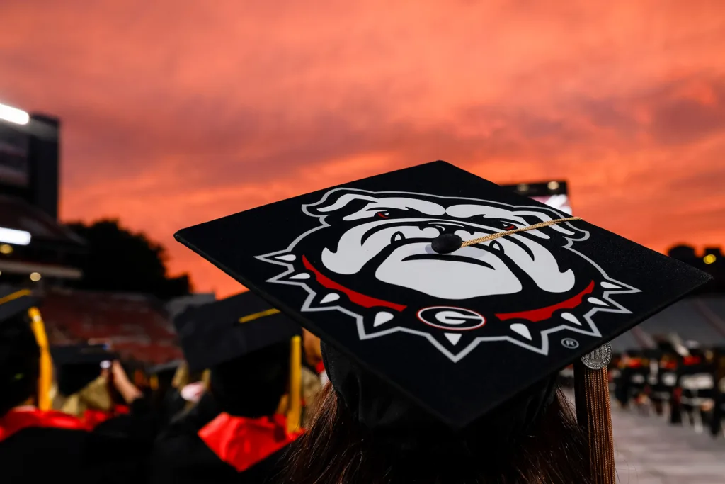 A graduation cap decorated with a large Georgia Bulldogs logo is silhouetted against a vivid orange sunset sky in the stadium.