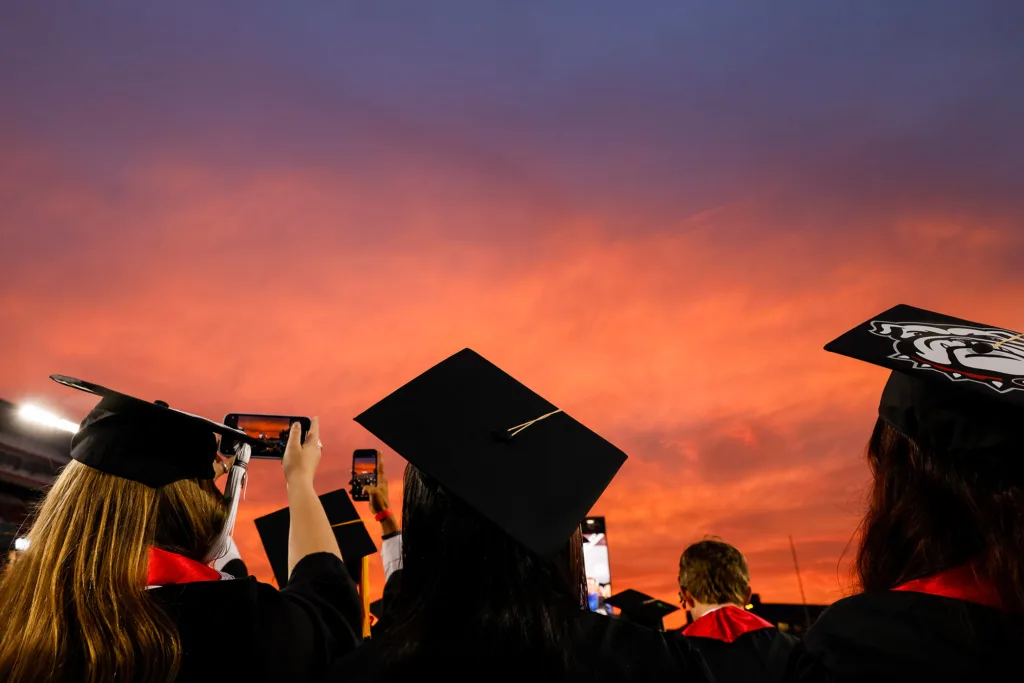 Graduates in caps and gowns hold up their phones to photograph the bright sunset sky during the ceremony.