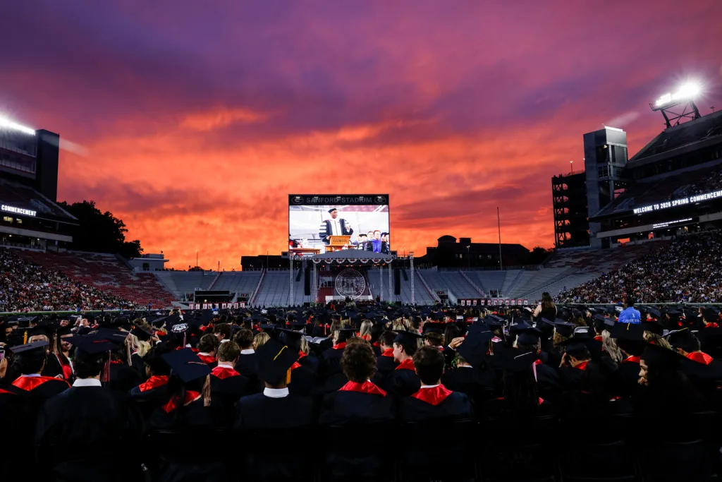 A wide view of Sanford Stadium shows rows of seated graduates facing the stage beneath a dramatic sunset.