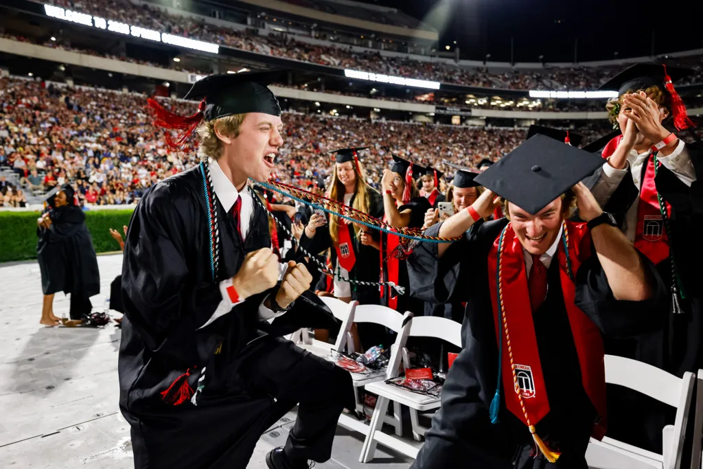 Two graduates celebrate energetically, cheering and laughing among a crowd of classmates.