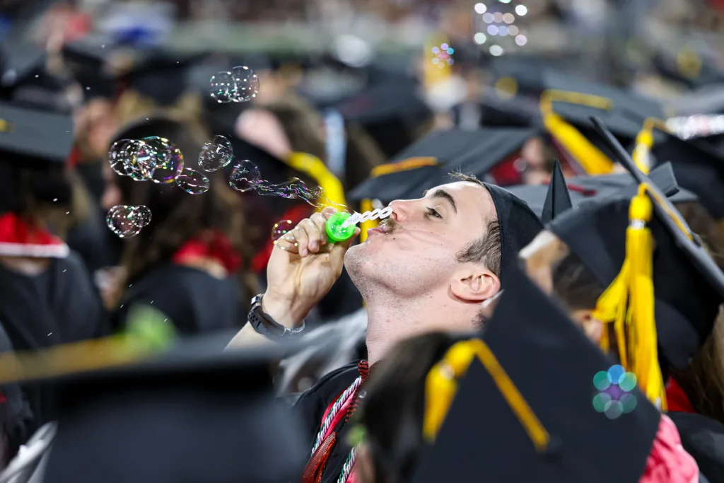 A graduate tilts their head back to blow bubbles that float above a sea of caps and tassels.
