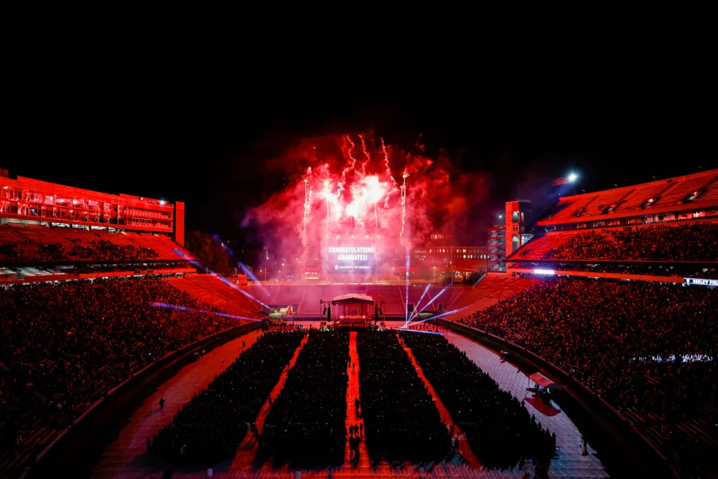 A wide nighttime view of Sanford Stadium shows red fireworks exploding behind the stage as a large screen reads “Congratulations, Graduates!”