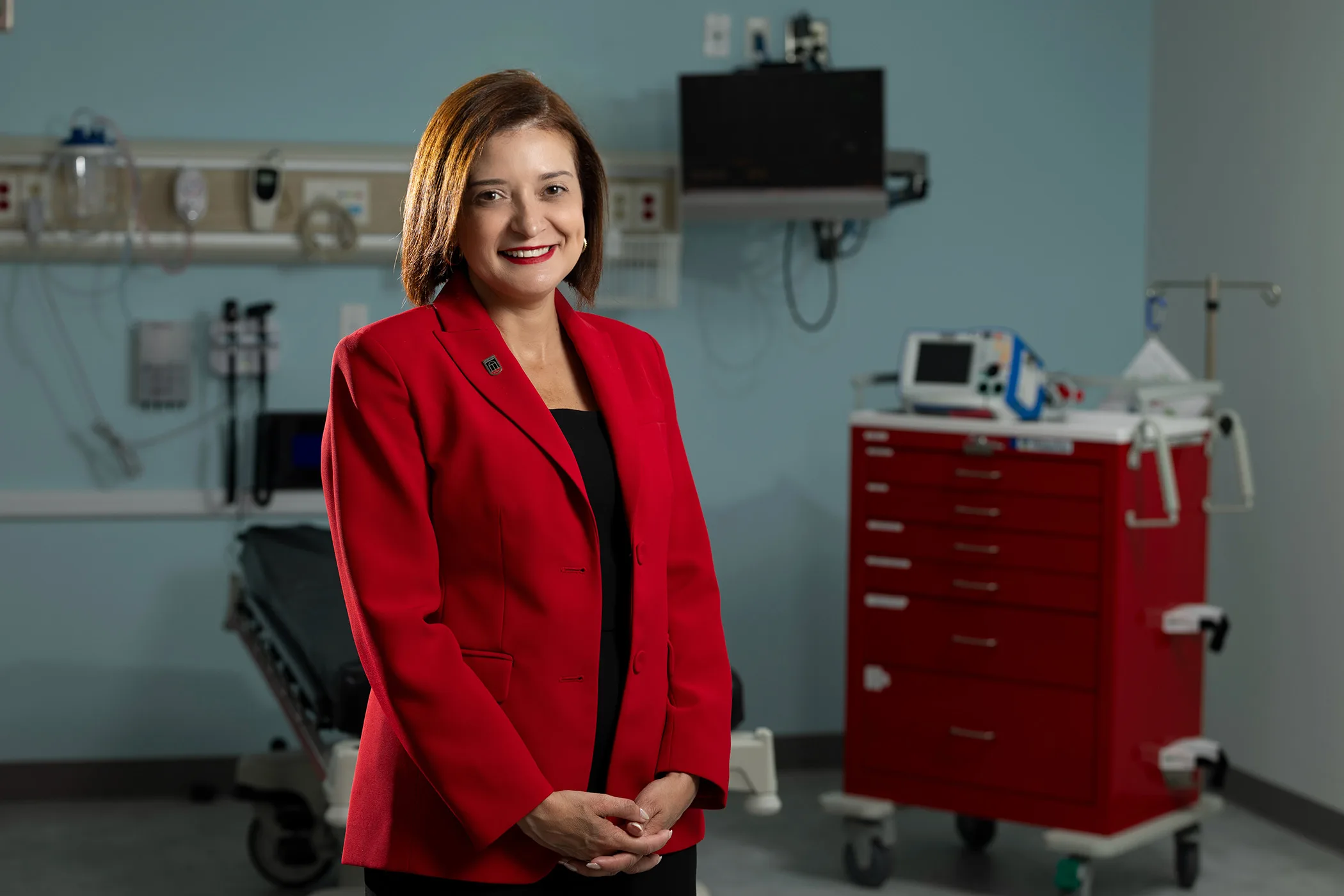 Laika Steiger in a red blazer poses in a medical simulation room with exam equipment behind her.