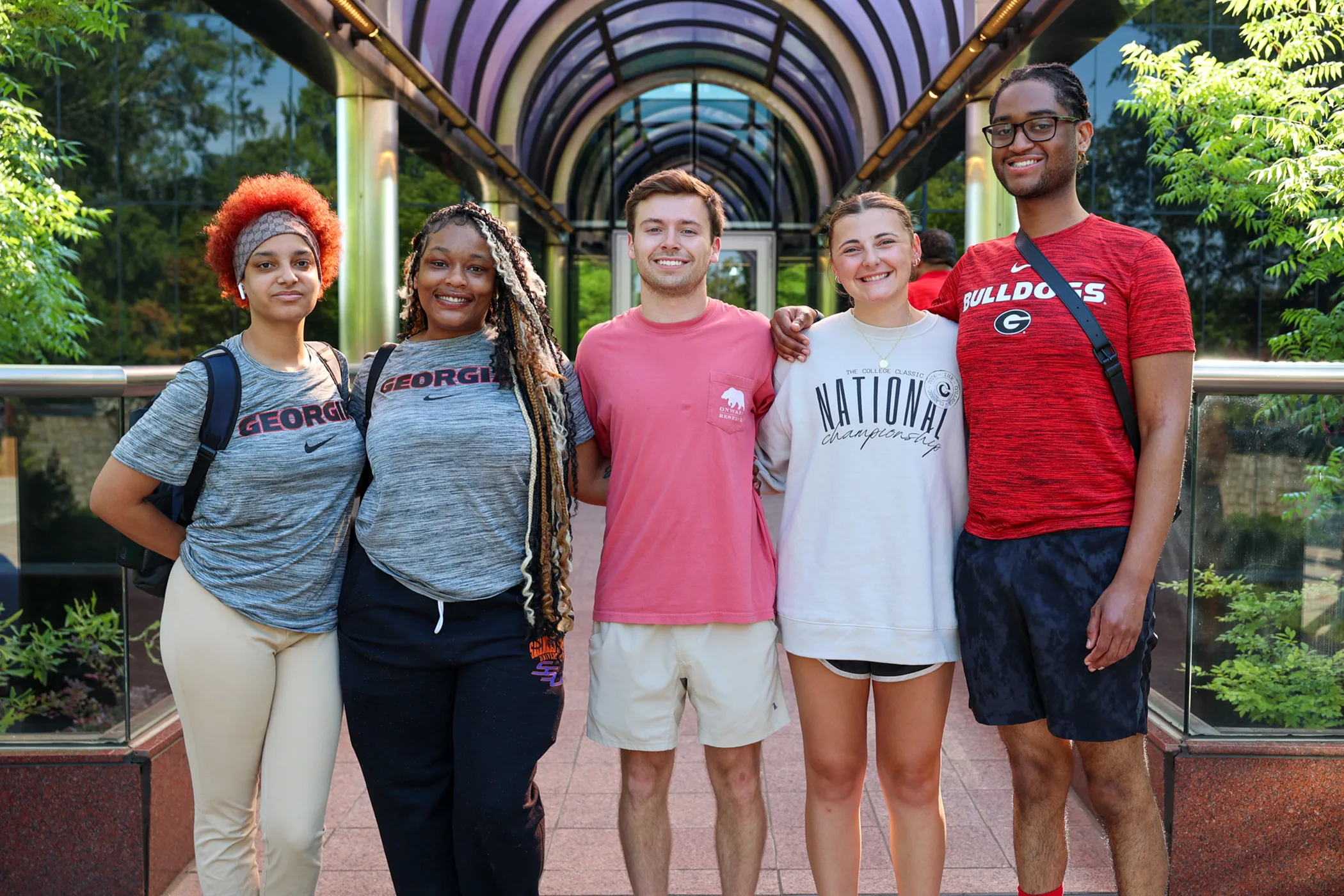 Five smiling students stand arm-in-arm beneath a covered walkway on campus.