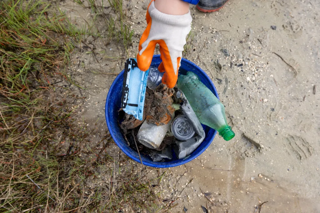 A gloved hand drops trash into a collection bucket on the sand in a coastal area.
