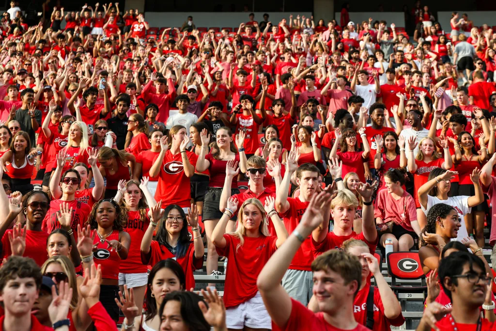 A large crowd of people in the stands of a football stadium. Most of them are wearing red shirts and raising their hands in the air.
