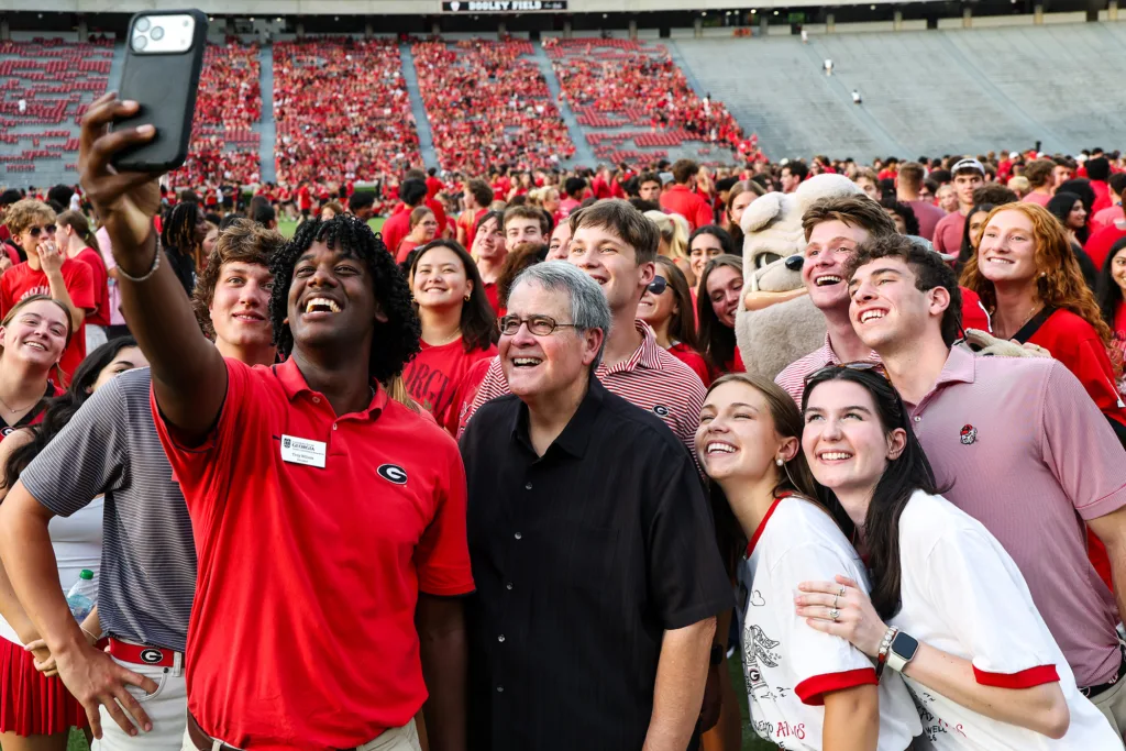 A young man in a red college polo takes a selfie of himself and a large group of people dressed in red, black, white and gray clothing.