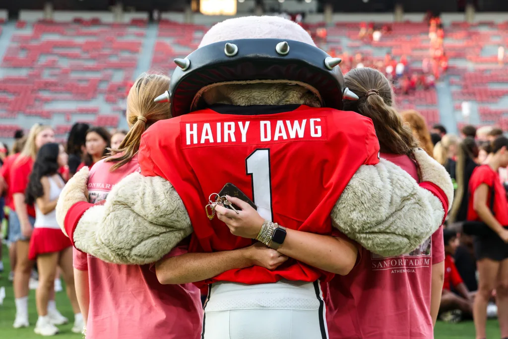 The back view of a large bulldog mascot hugging a young woman on each side of him. The mascot is wearing a red jersey with the number “1” and the words “Hairy Dawg.”