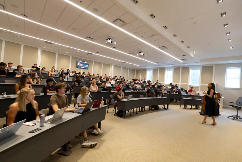 A female professor talks to a large group of college students seated in a lecture hall.