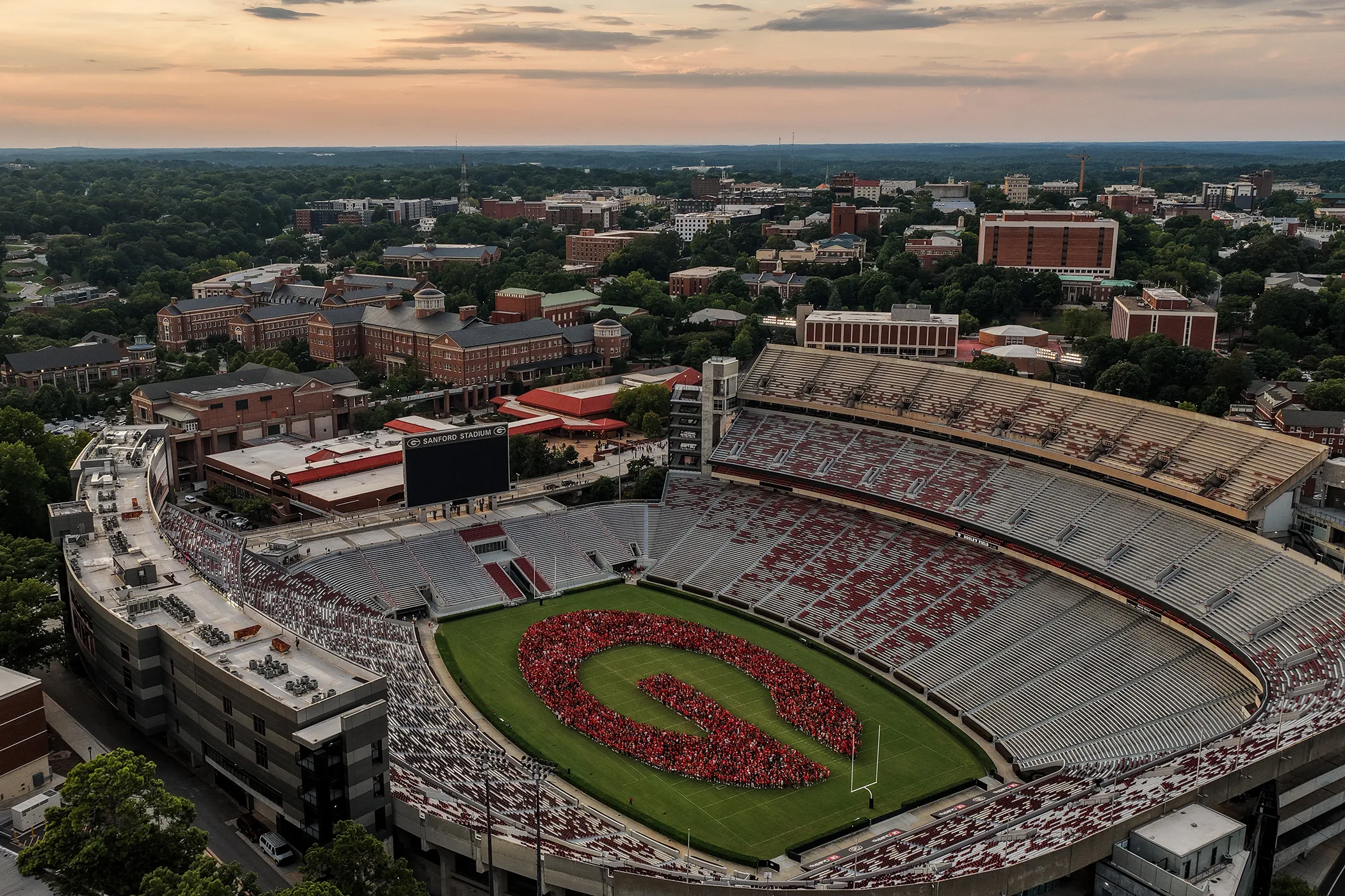 An aerial view shows students in red forming the shape of Georgia's "Power G" logo on the field of Sanford Stadium, with the campus and skyline stretching into the distance.