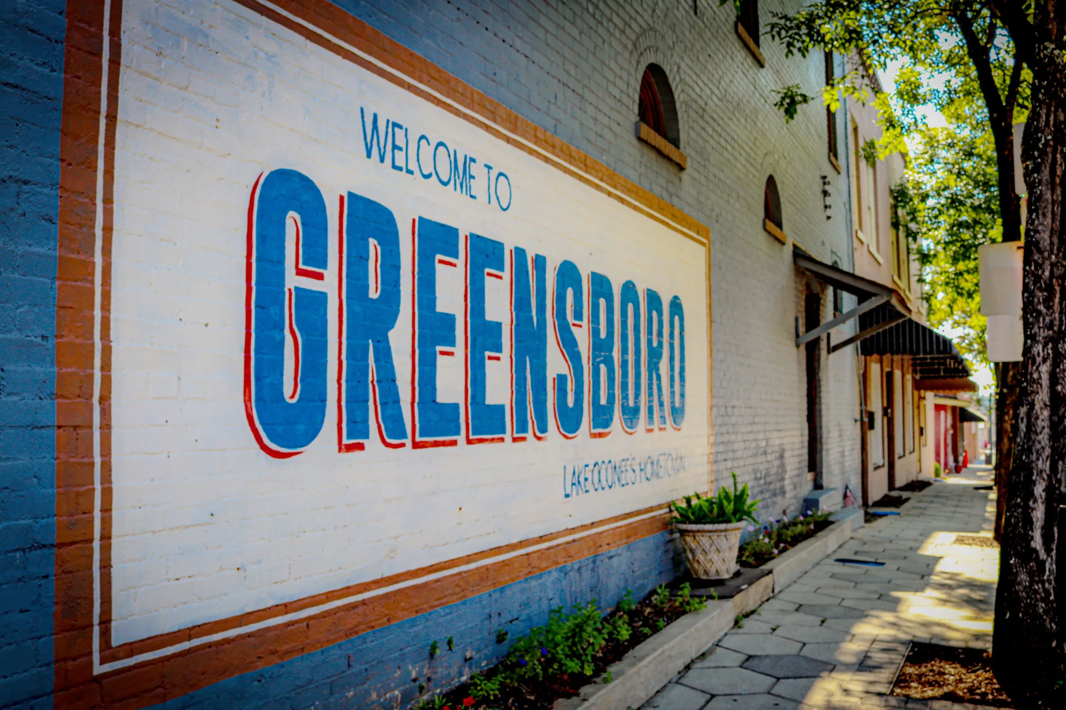 A large mural reading "Welcome to Greensboro, Lake Oconee's Hometown" is painted on the brick wall of a downtown building along a shaded sidewalk.
