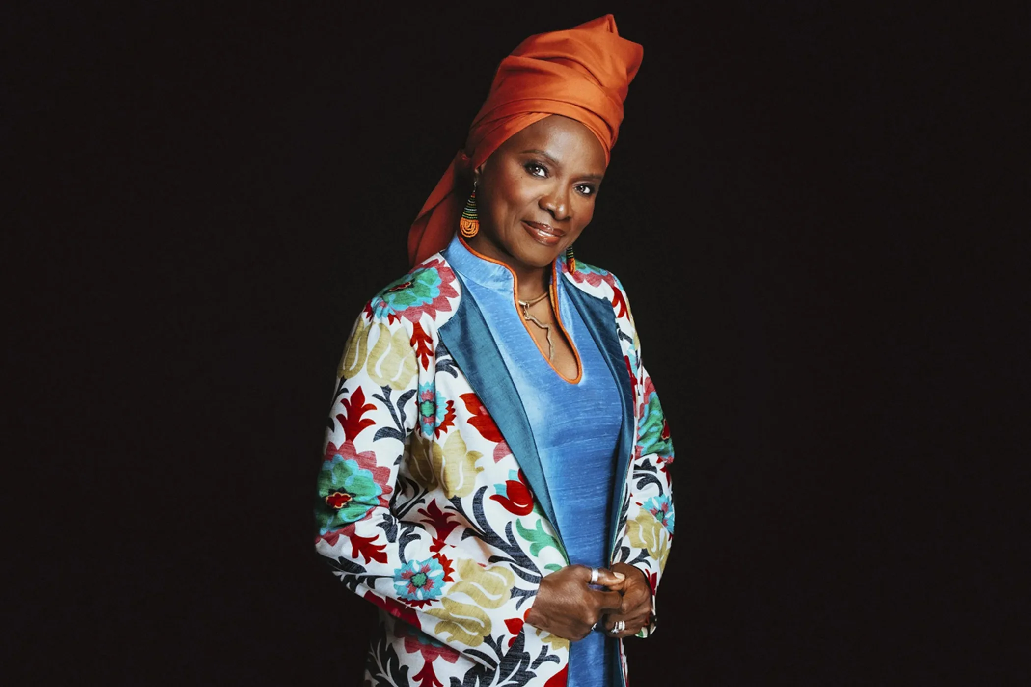 Angélique Kidjo wears an orange head wrap, beaded earrings and a floral jacket over a blue top against a black background.
