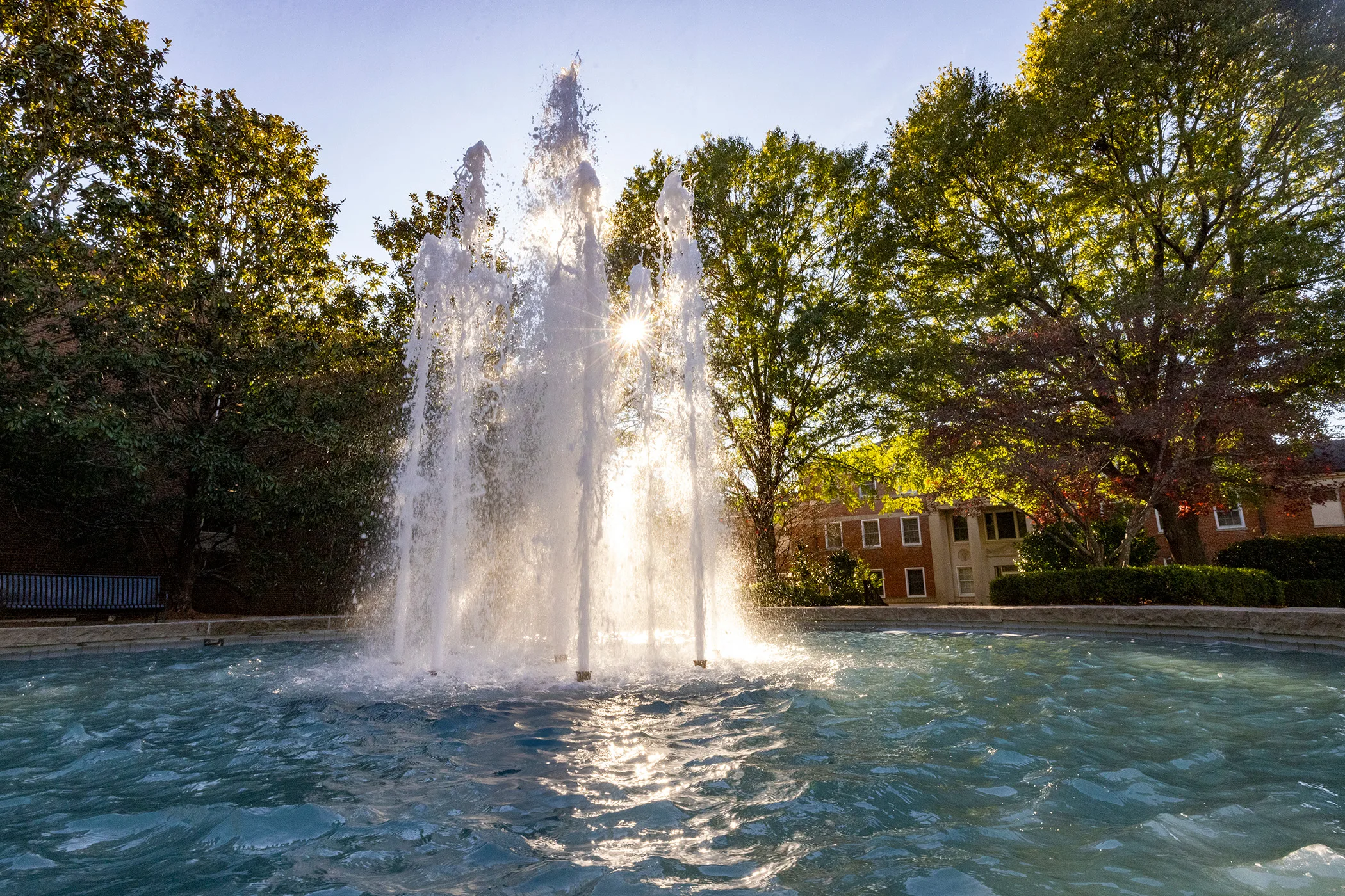 Sunlight glints through the spray of a campus fountain, backlit against a canopy of trees and a brick building.