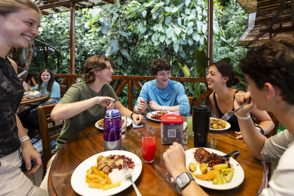 A group of people laugh and enjoy dinner at a restaurant with wide glass windows that show a leafy jungle outside.