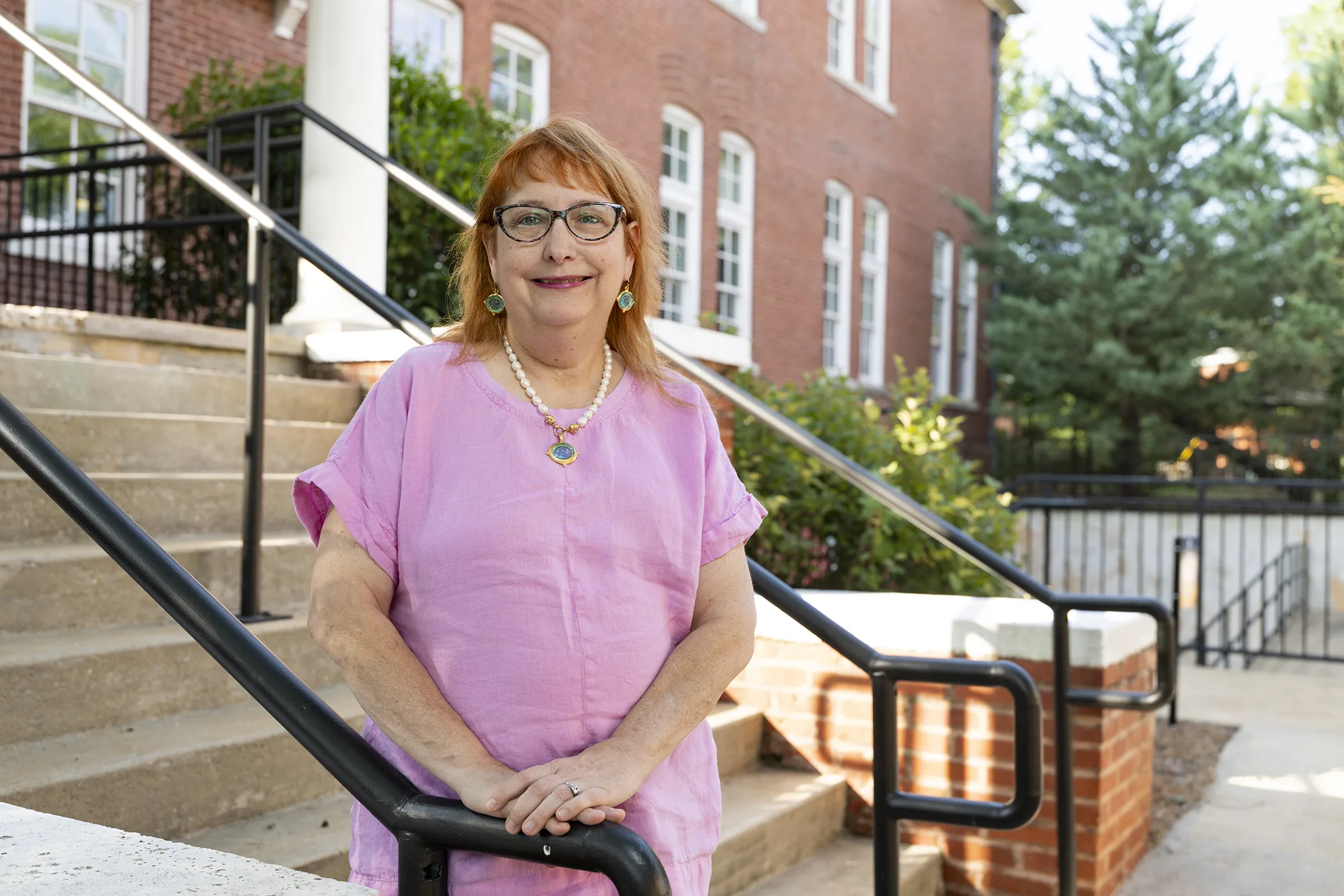 A woman wearing pink stands on the steps of an academic building, smiling toward the camera