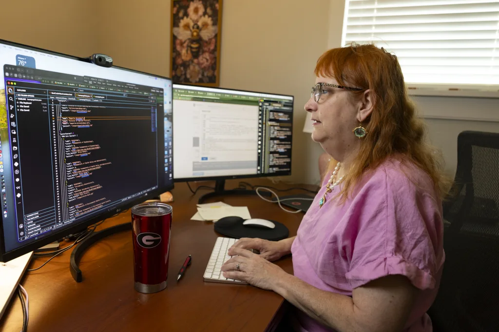 A woman sits at a desk with two computer screens, working on coding
