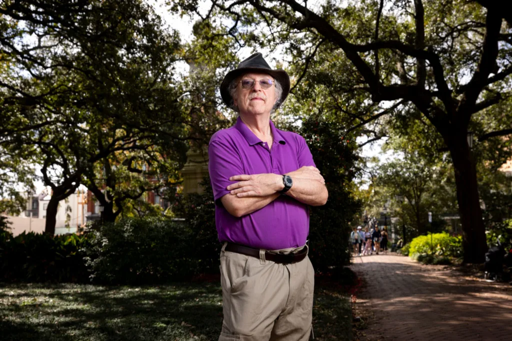A man standing under trees in Savannah wearing a hat and sunglasses while crossing his arms.